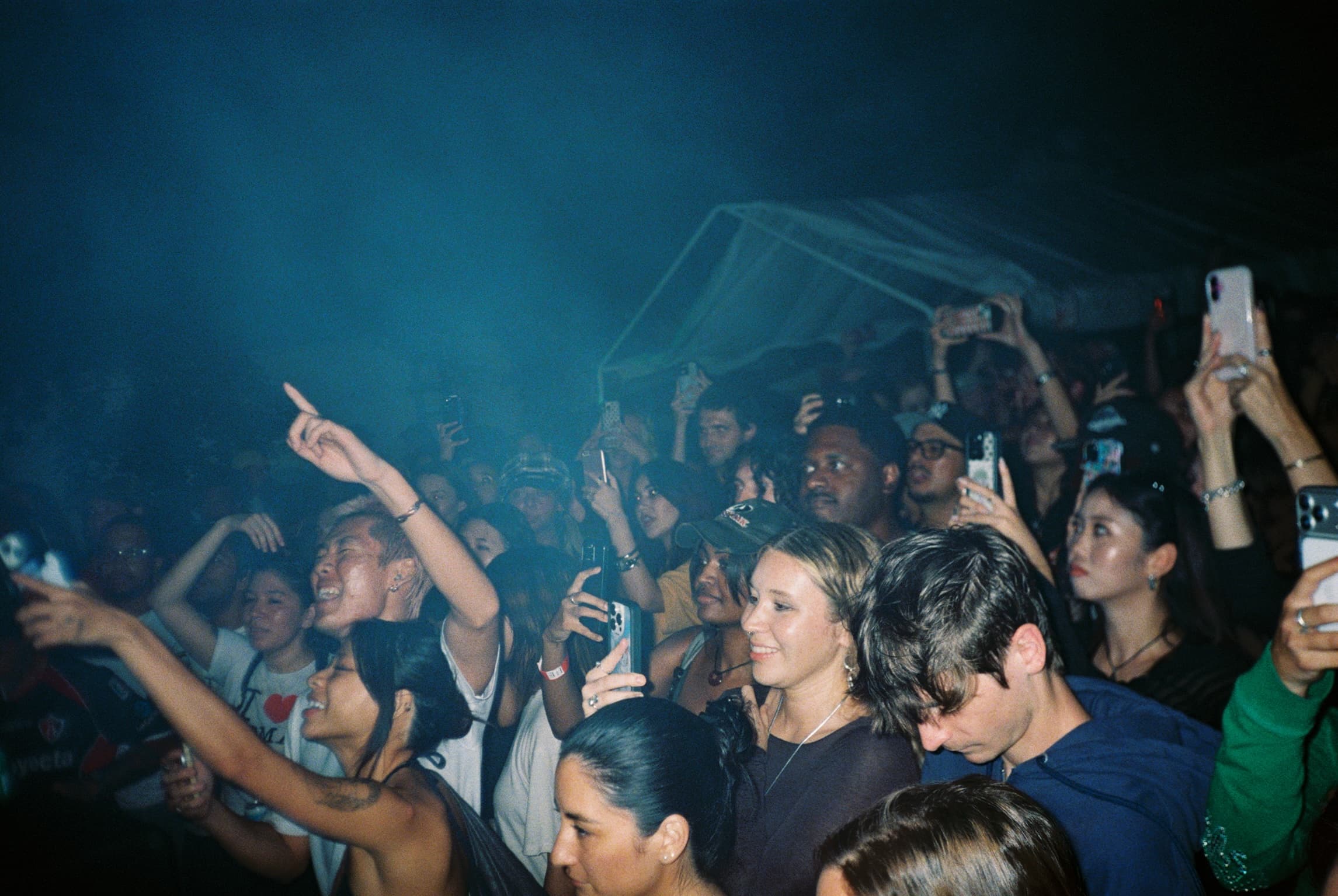 A packed crowd at a show, people singing, pointing at the stage and holding their phones up to film.
