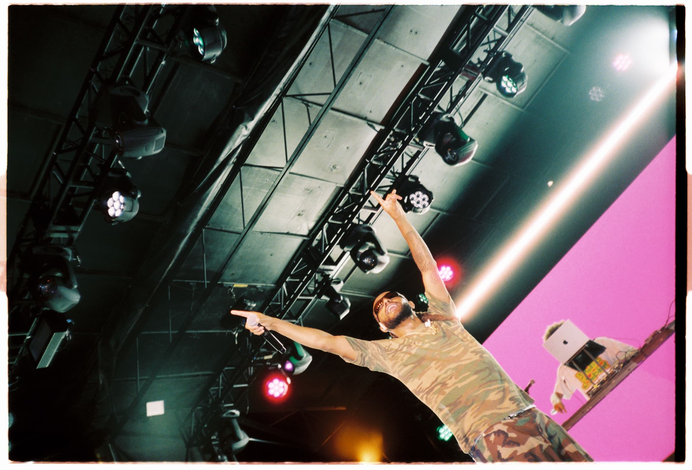 A performer on stage pointing up at the lighting rig, a pink screen behind him.