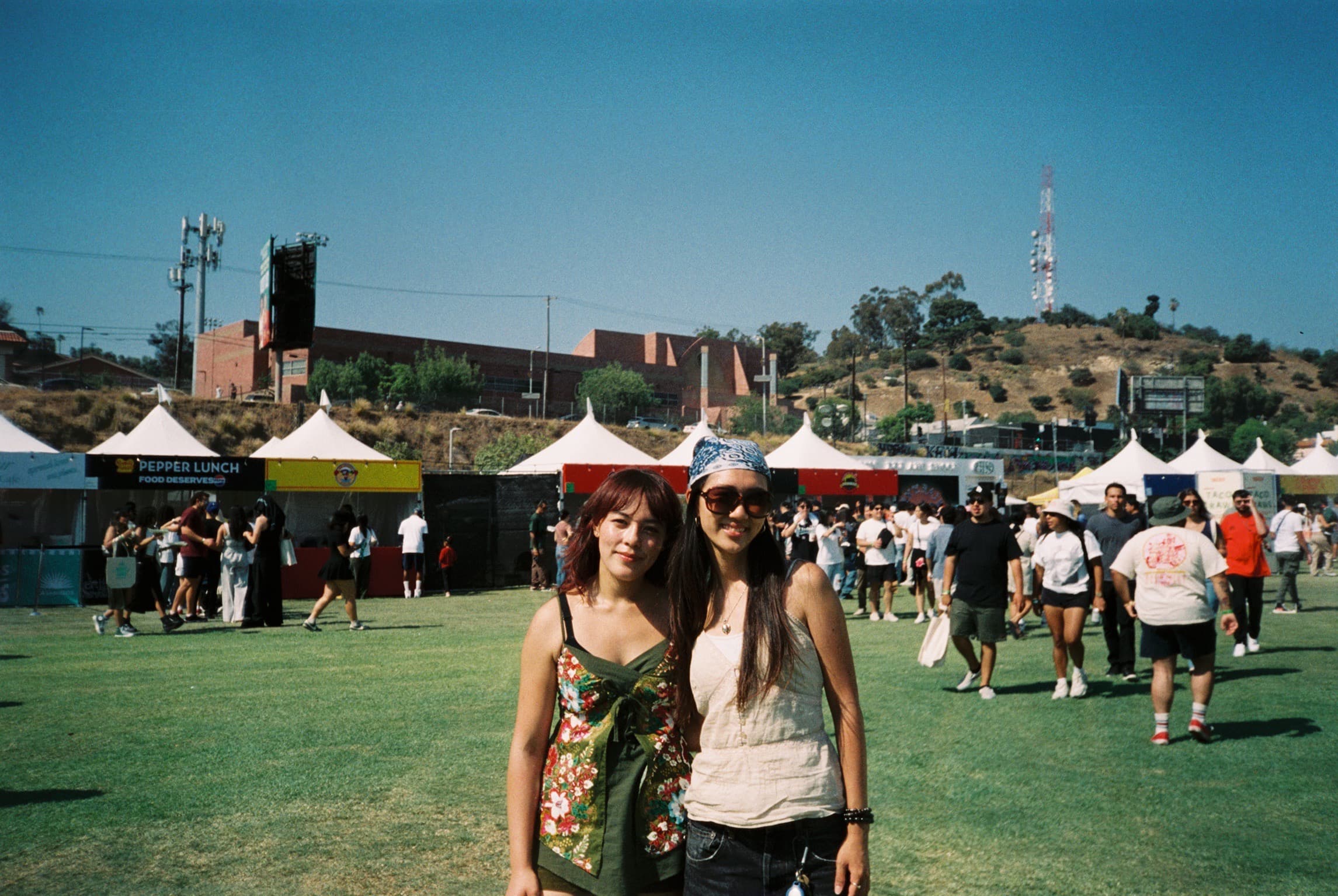 Two friends posing together on the grass at a daytime festival.